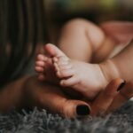 Infant's feet being held by a woman's hand with painted and manicured hands resting on a gray blanket